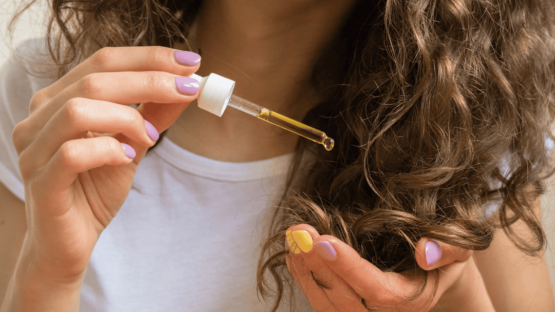 Kvinna applicerar hårolja i håret. Man ser endast händerna och håret som är krulligt. Naglarna har nagellack i olika färger. 