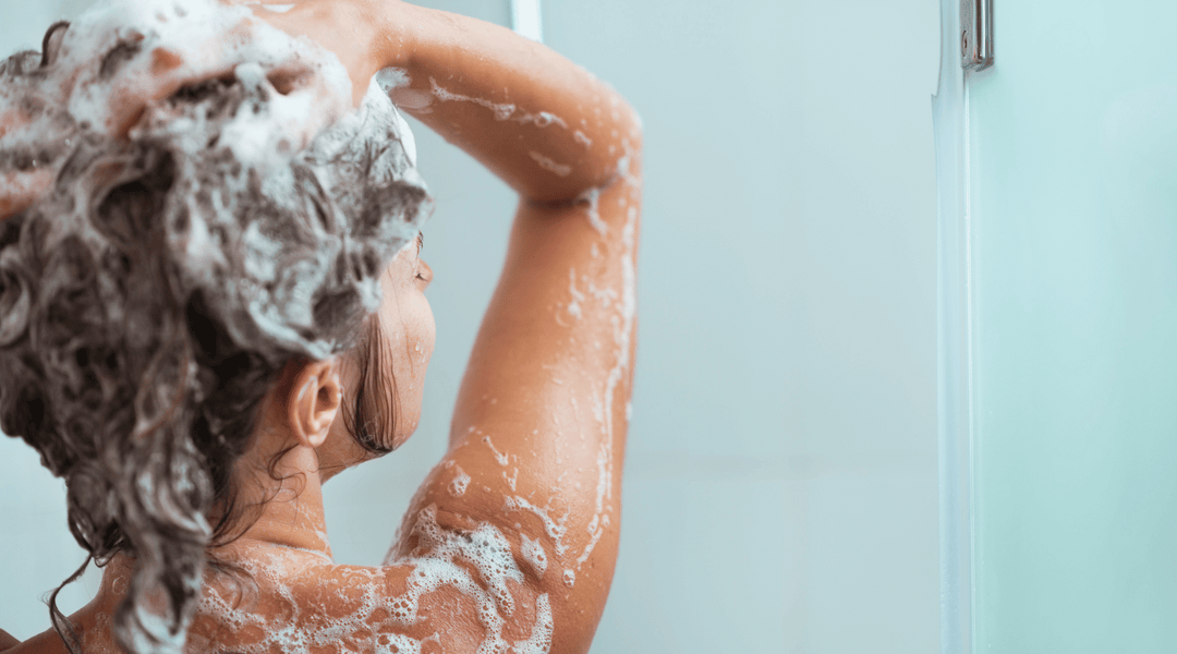 Person tvättar håret, täckt av schampo, i dusch, sedd bakifrån med upplyfta armar och lockigt hår.