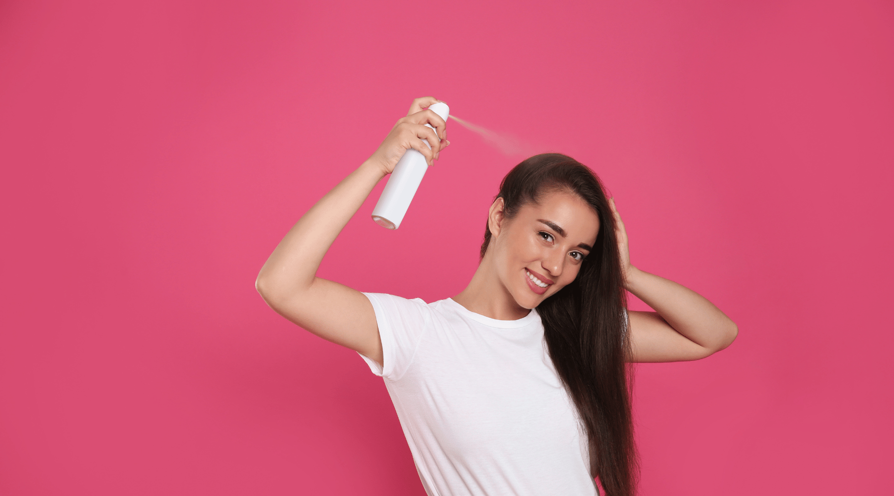 Leende kvinna med långt hår sprayar hårprodukt, rosa bakgrund.