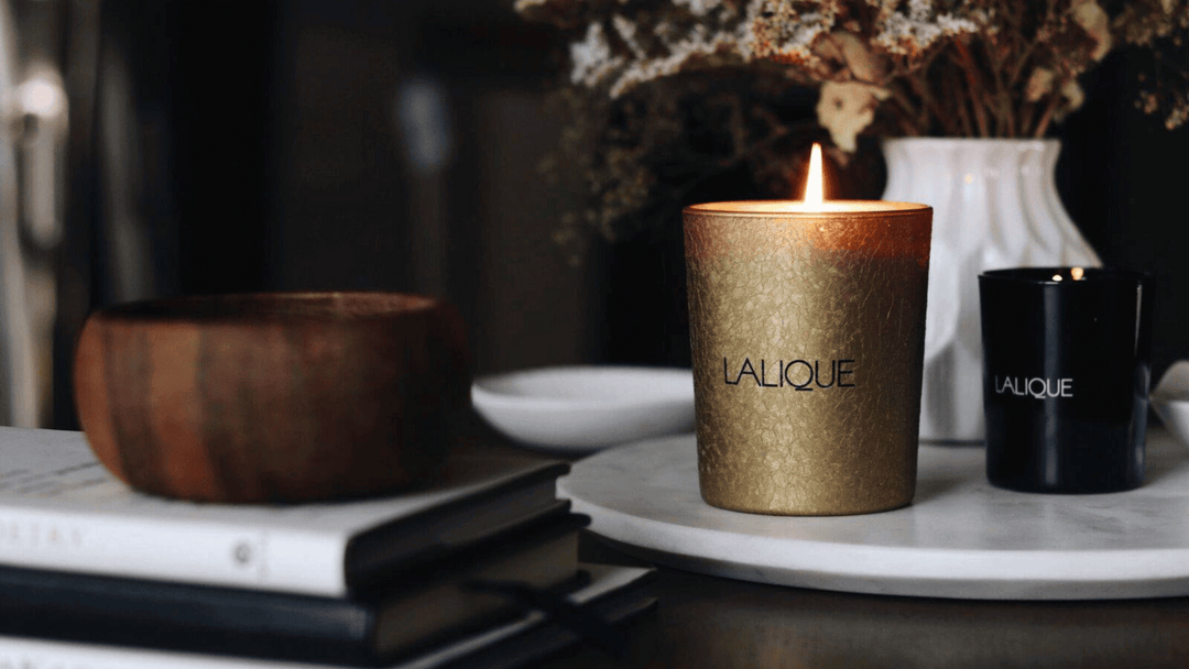 Ett tänt guldigt doftljus märkt "LALIQUE" på en marmorbord, med ett släckt svart ljus bakom och en bokstapel intill.