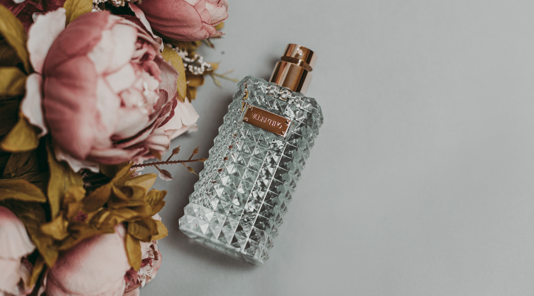 En detaljerad VALENTINO parfymflaska bredvid en bukett av blekrosa pioner, vilket skapar en känsla av lyx och romantik.