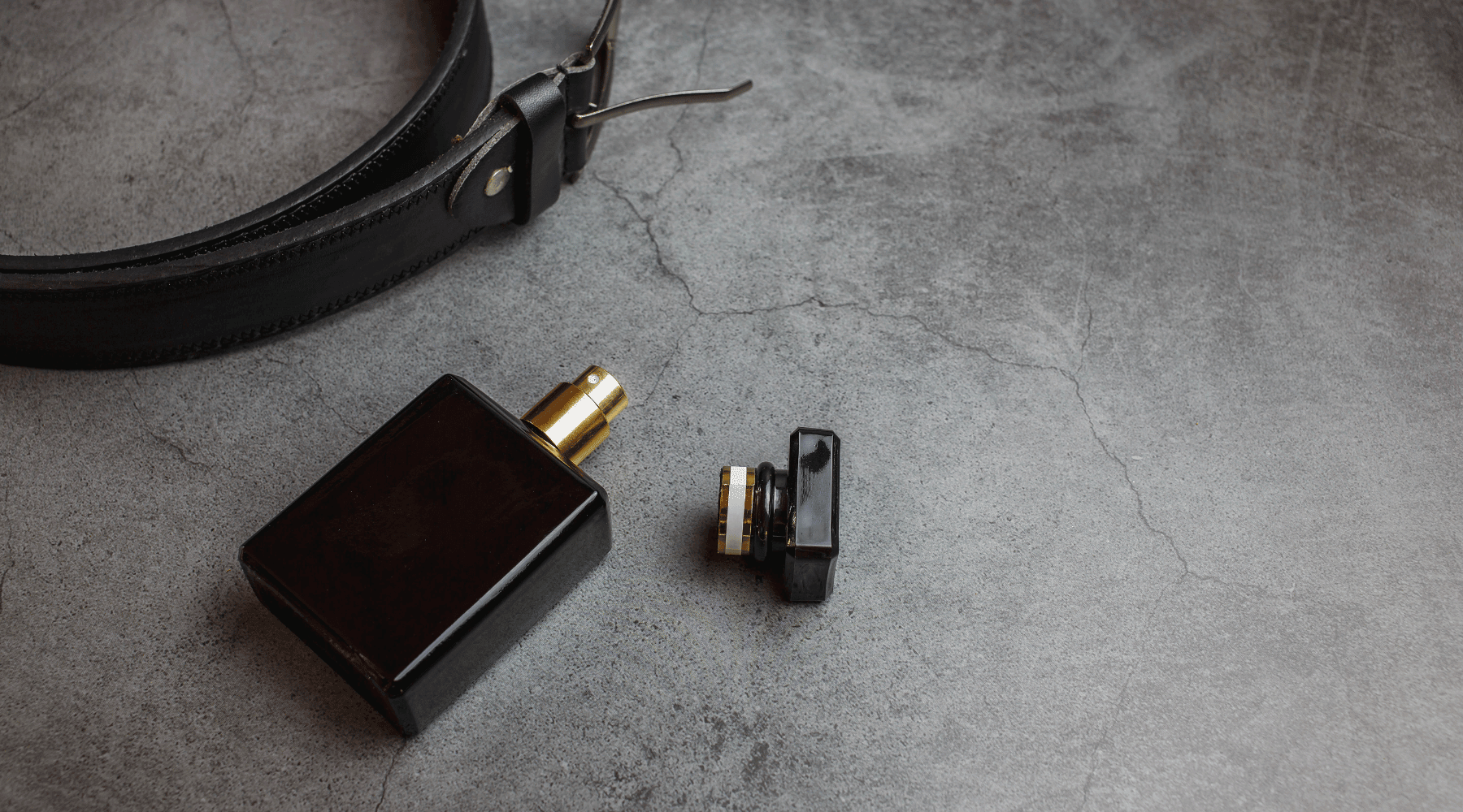 Ett svart bälte och en mörk parfymflaska med gulddetaljer på grå bakgrund.