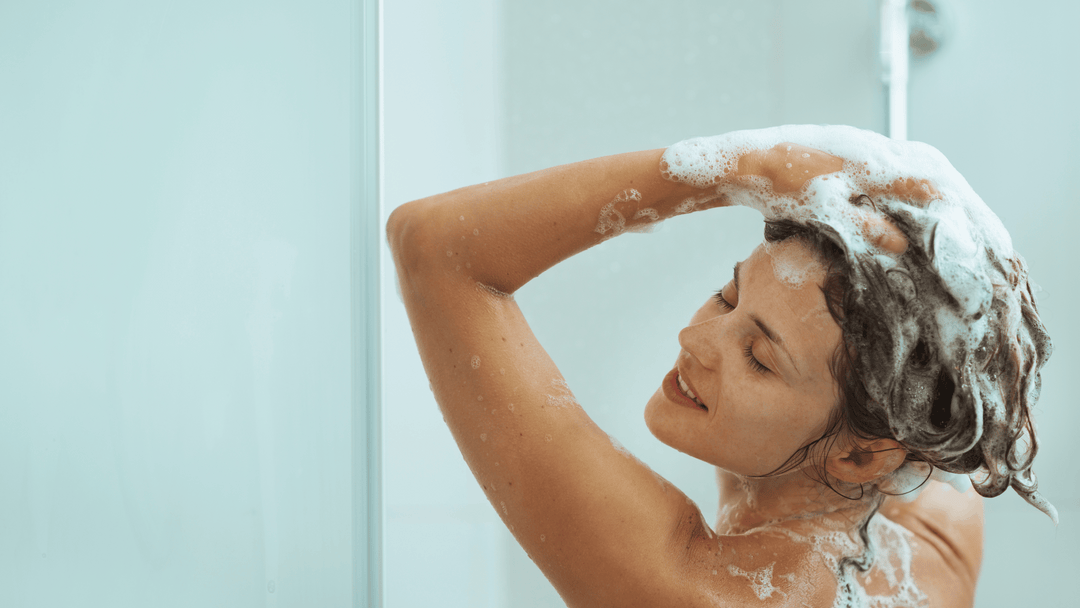 Kvinna tvättar håret, täckt av skum, i dusch, synlig lycka.